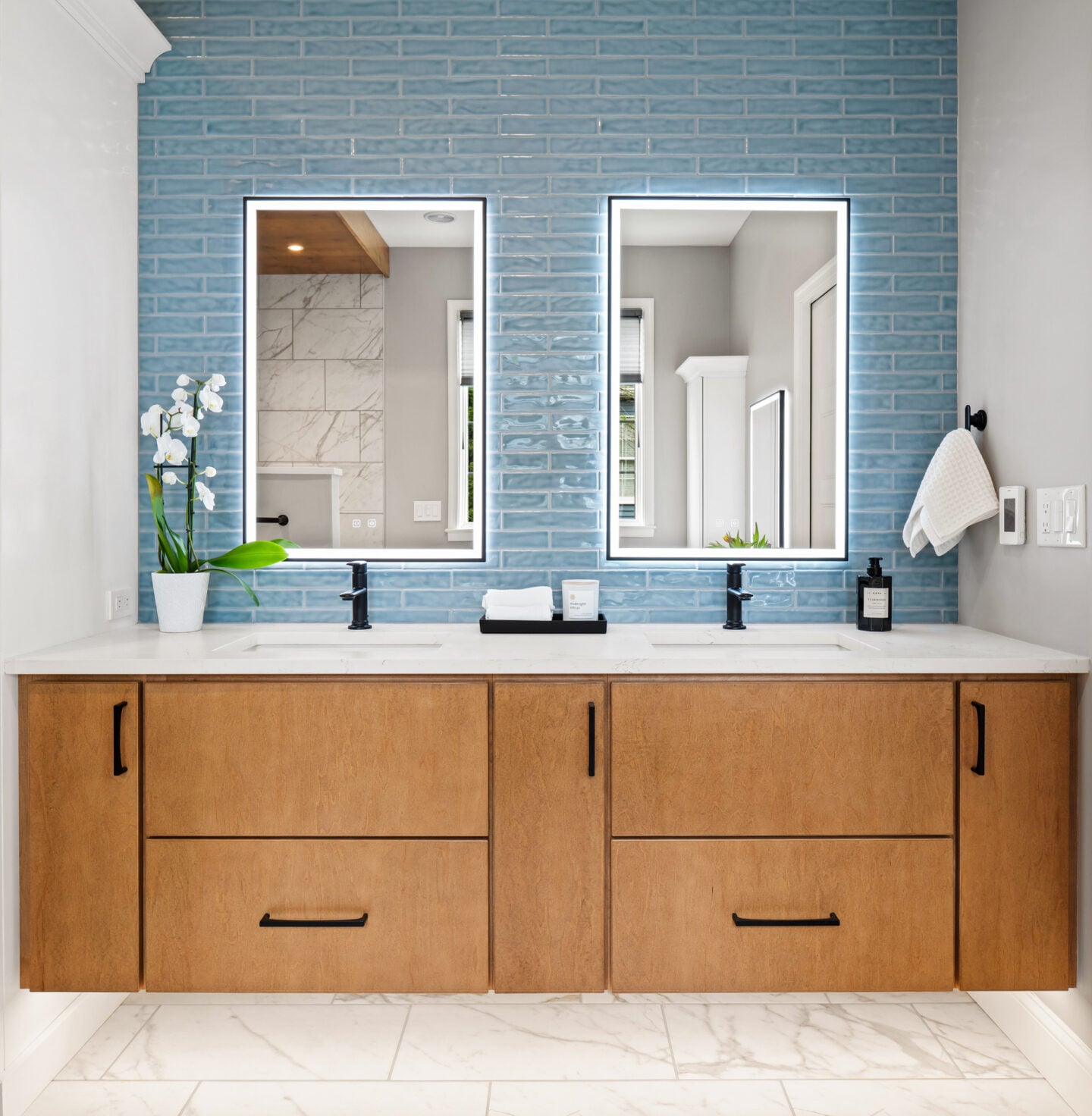 Modern floating bathroom vanity with warm maple cabinetry, quartz countertop, and illuminated mirrors against glossy blue tile.