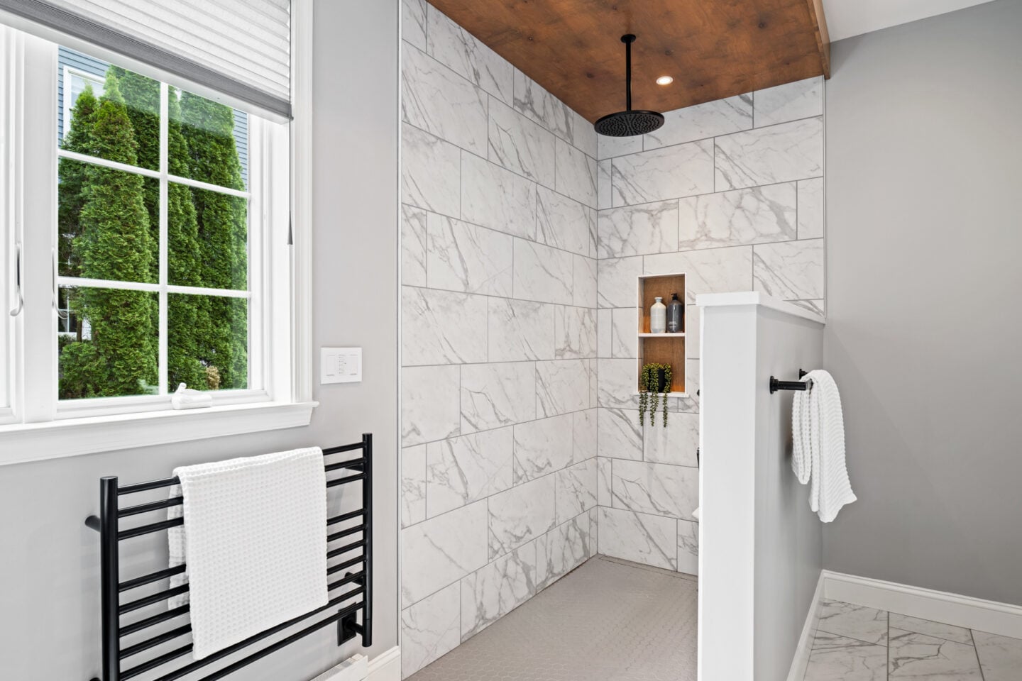 Bright walk-in shower with porcelain tile walls, wood soffit ceiling detail, black fixtures, and natural light from nearby window.