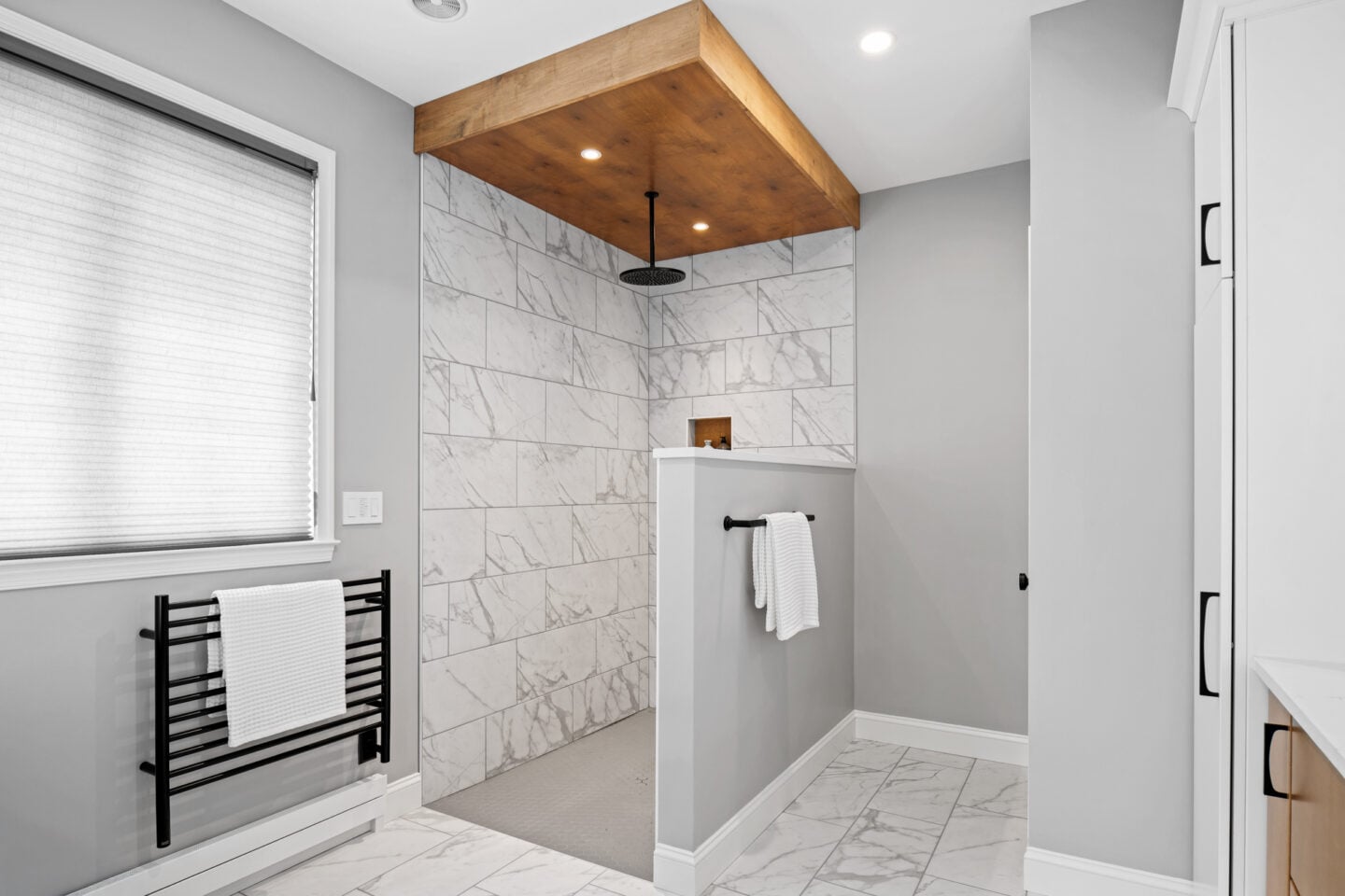 Walk-in shower with stained wood soffit, marble-look porcelain tile, and rain shower head in a modern Doylestown primary bathroom remodel.